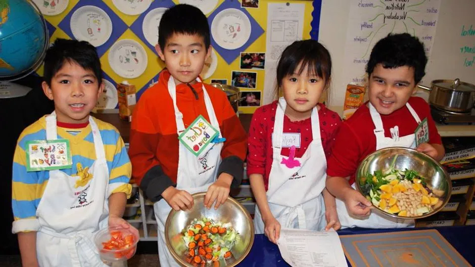 children wearing aprons in class holding bowls of food they prepared